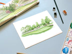 golf landscape from practice round watercolor archival print laying on a desk with a sketchbook
