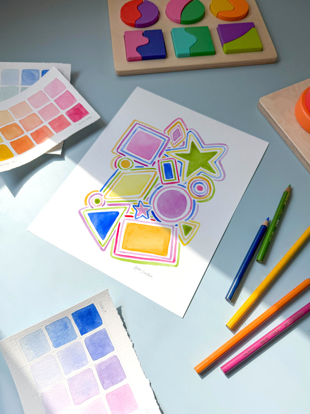 Colorful geometric shapes on a piece of paper surrounded with coloring books and pencils on a light blue background