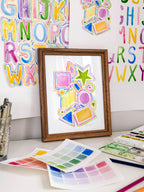 Colorful geometric shapes archival watercolor print in a wooden frame on a desk with art supplies and alphabet letters in the background.
