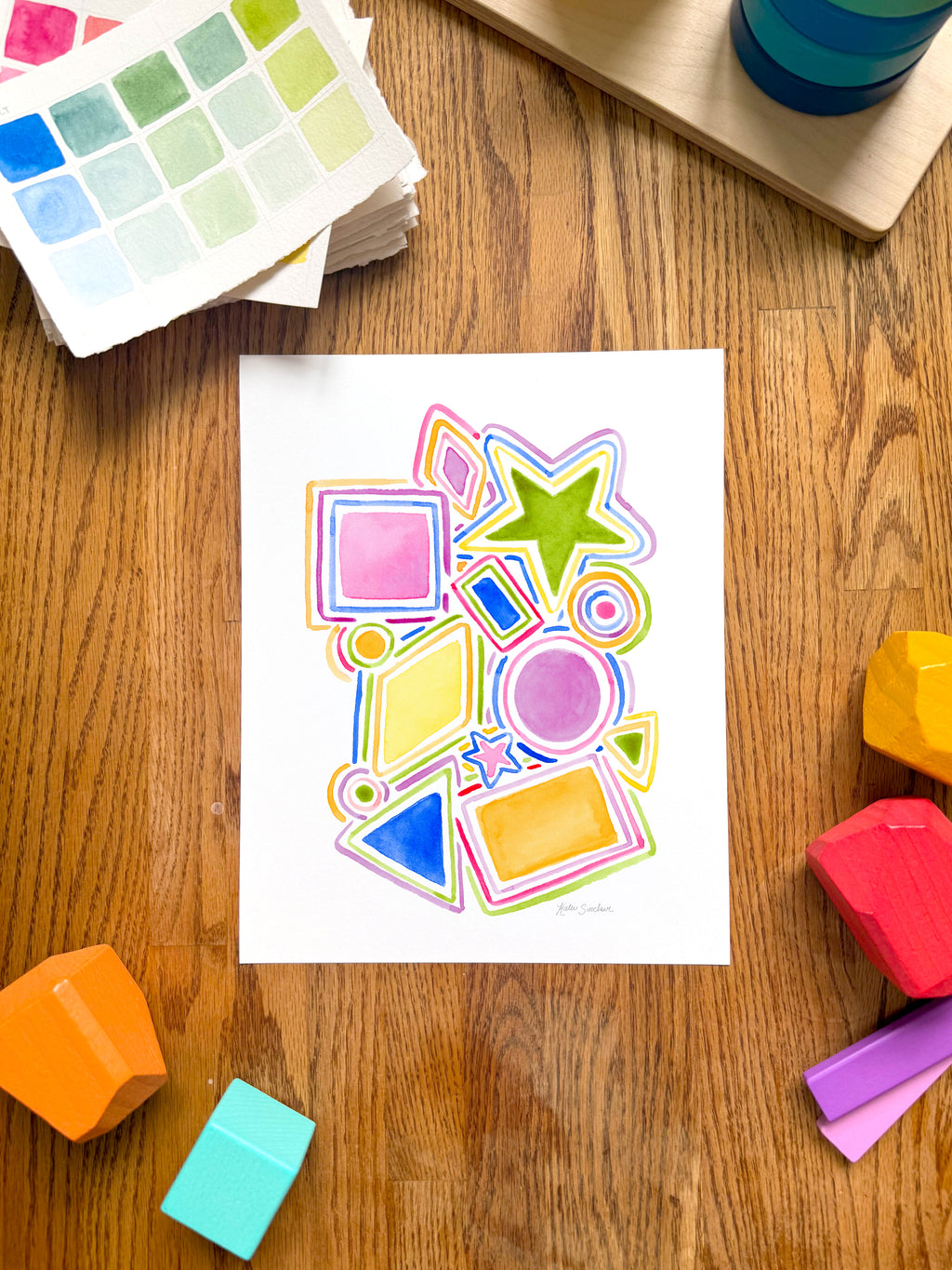 colorful watercolor shapes and lines archival print, surrounded by geometric toys on a wooden surface.  hand-painted by katie sinclair and perfect for a playroom or kid bedroom.