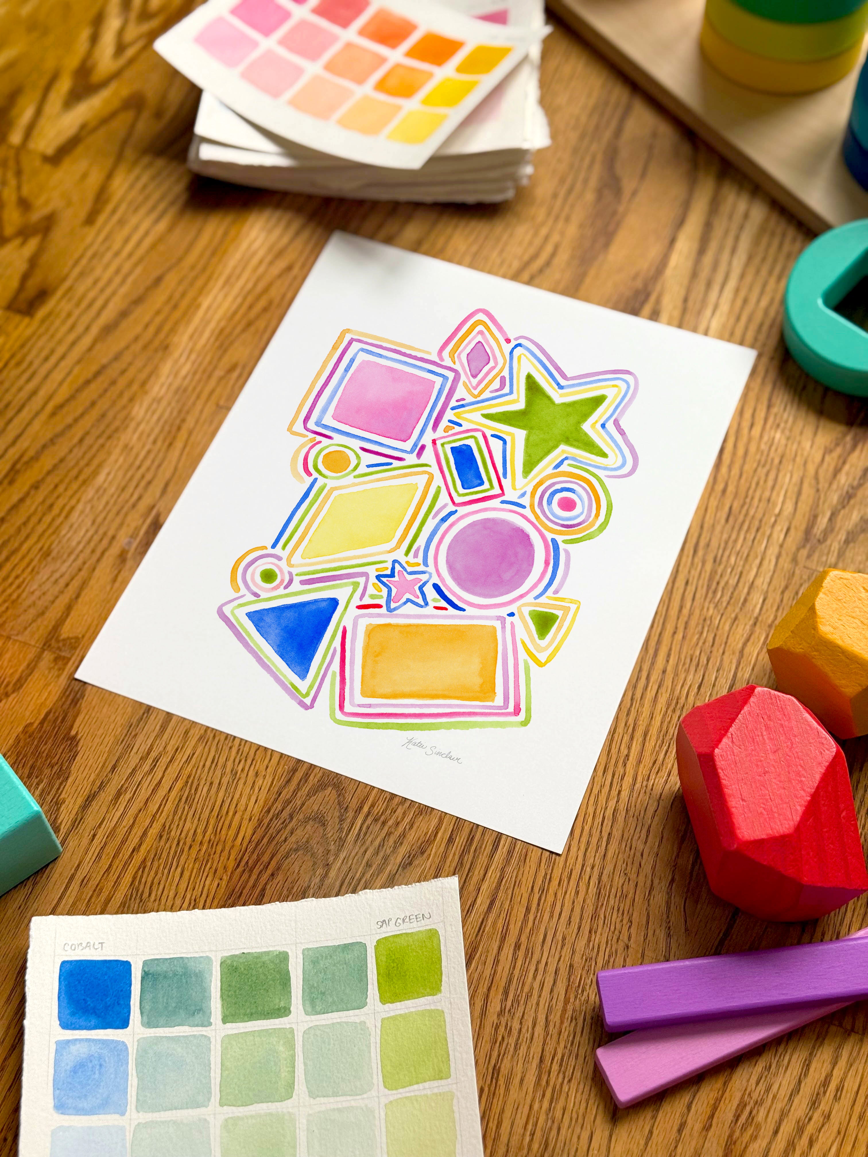 colorful watercolor shapes and lines archival print, surrounded by geometric toys on a wooden surface.  hand-painted by katie sinclair and perfect for a playroom or kid bedroom.