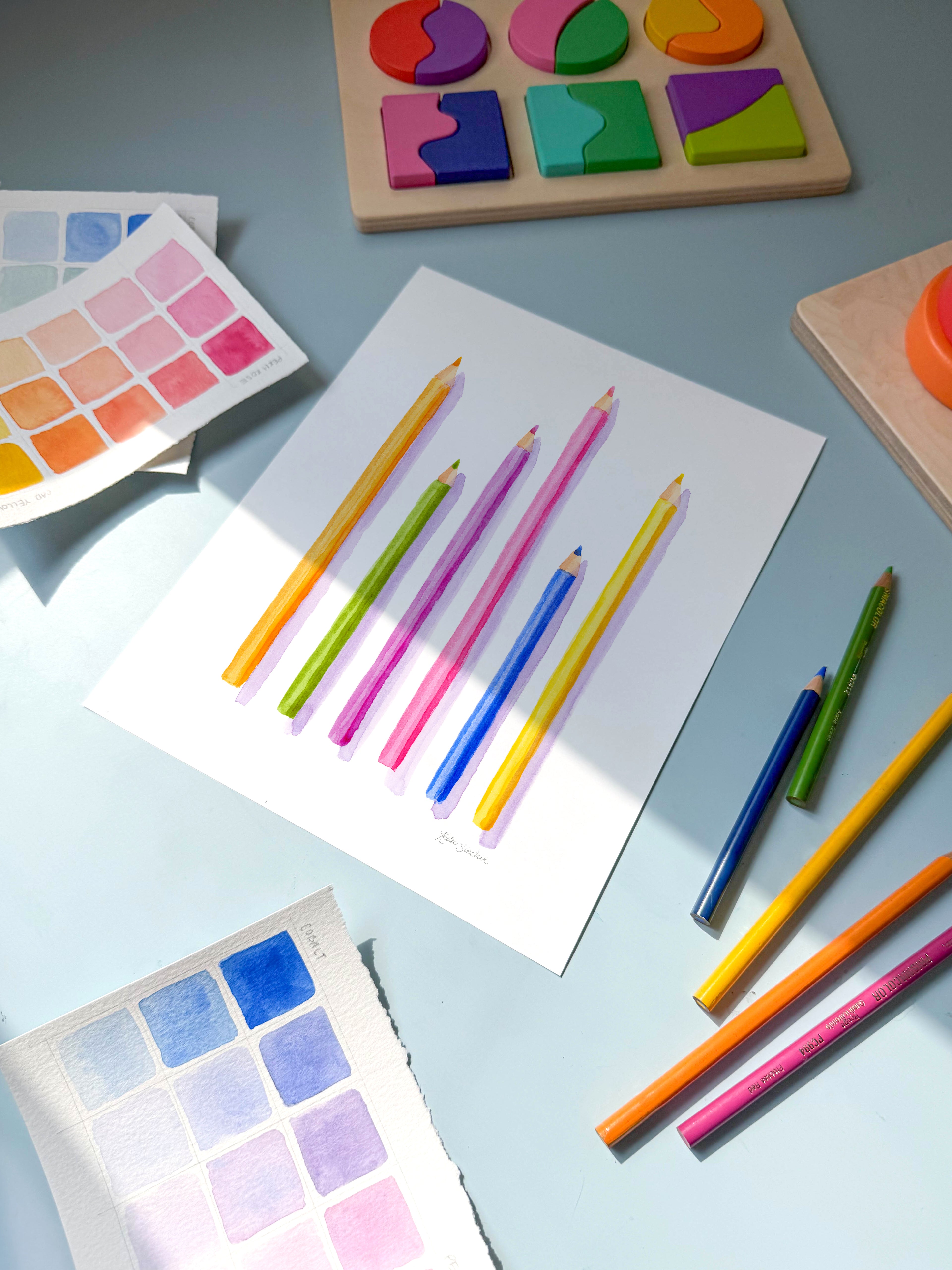 colored pencils watercolor archival print by Katie Sinclair on a desk with colored pencils and a wooden puzzle.