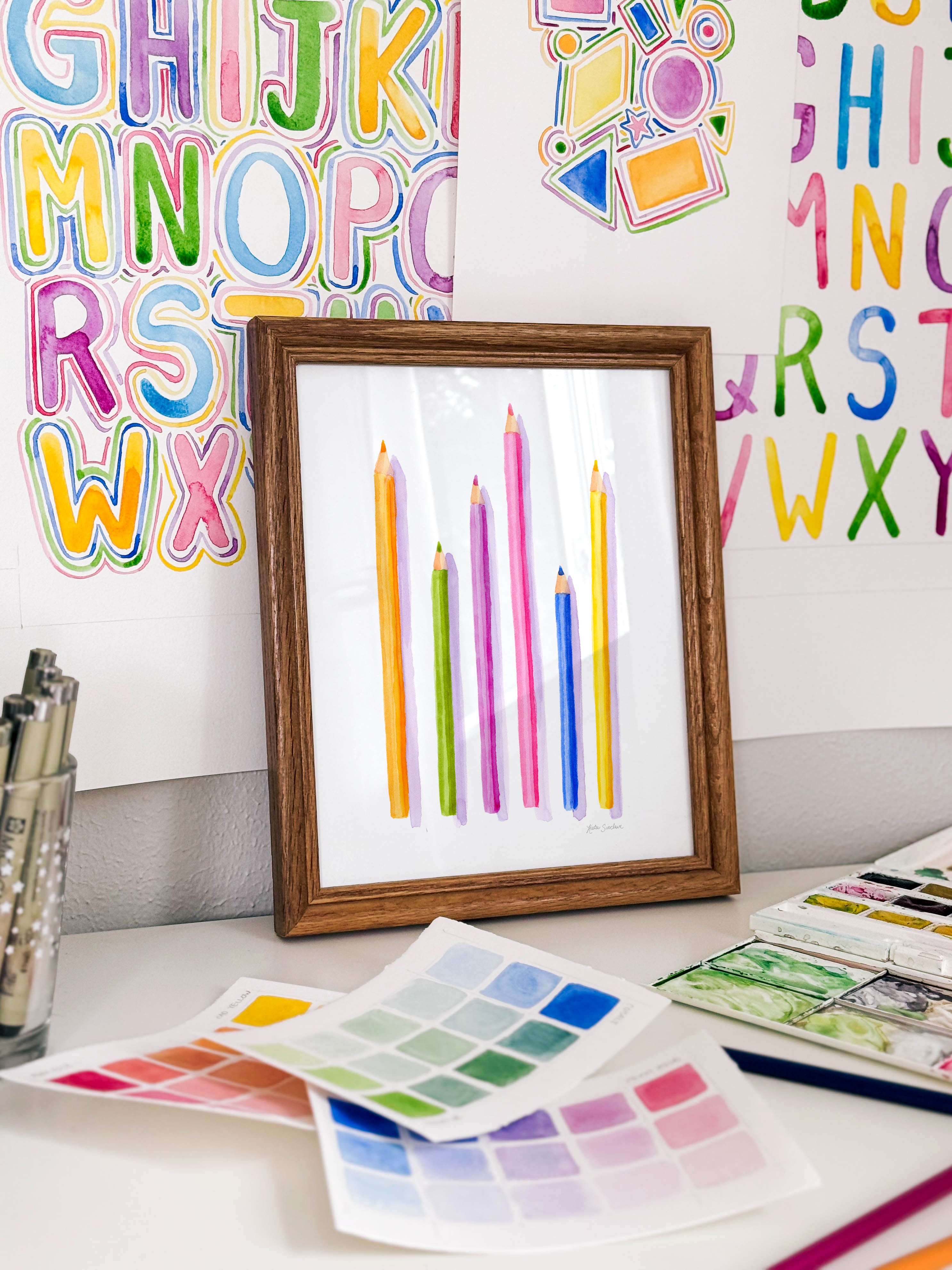 Framed watercolor pencils archival print by Katie Sinclair on a desk with colorful letters on the wall