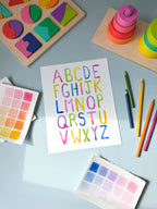 simple alphabet watercolor archival print by Katie Sinclair on a desk with colored pencils and a wooden puzzle.