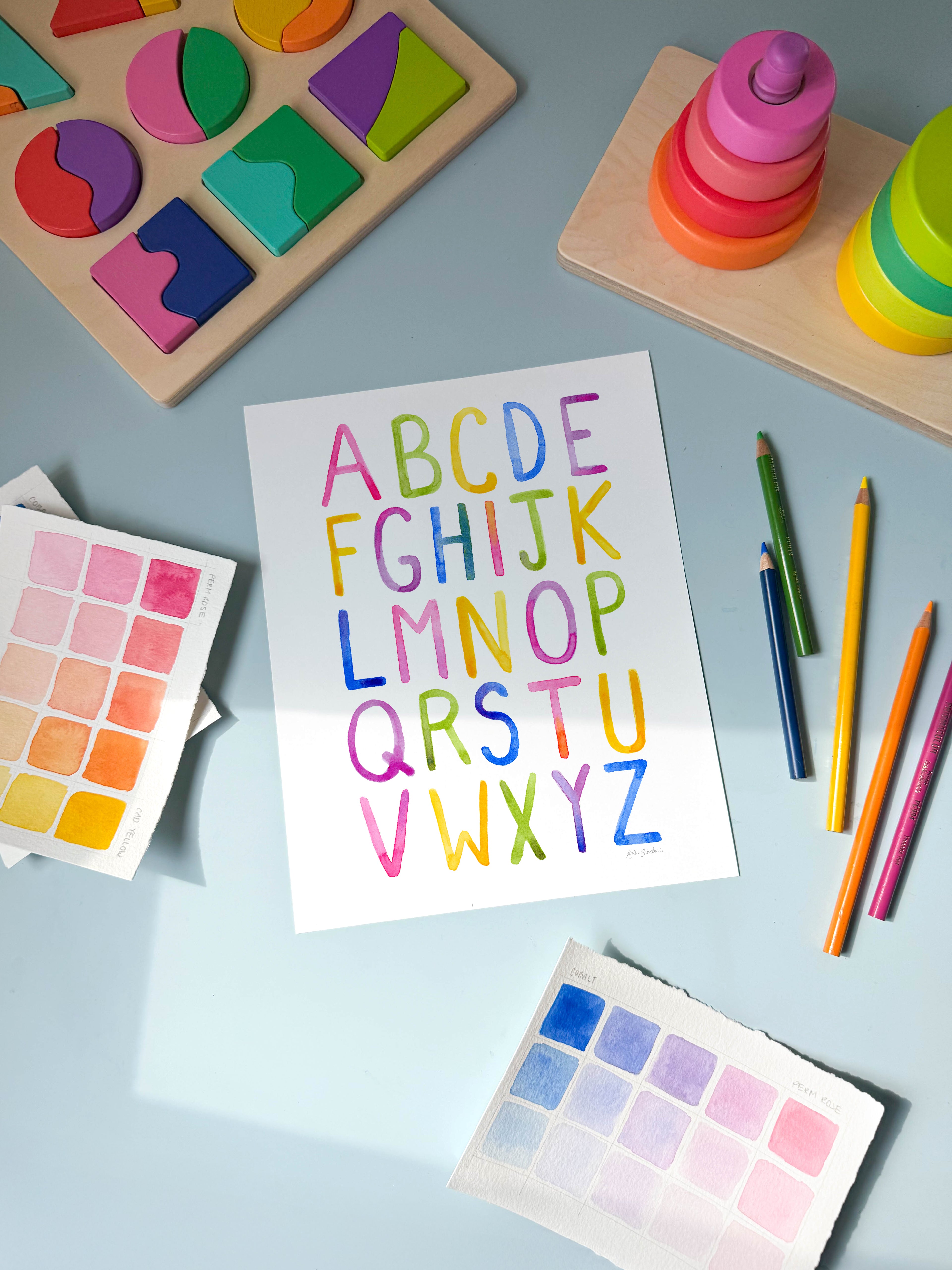 simple alphabet watercolor archival print by Katie Sinclair on a desk with colored pencils and a wooden puzzle.