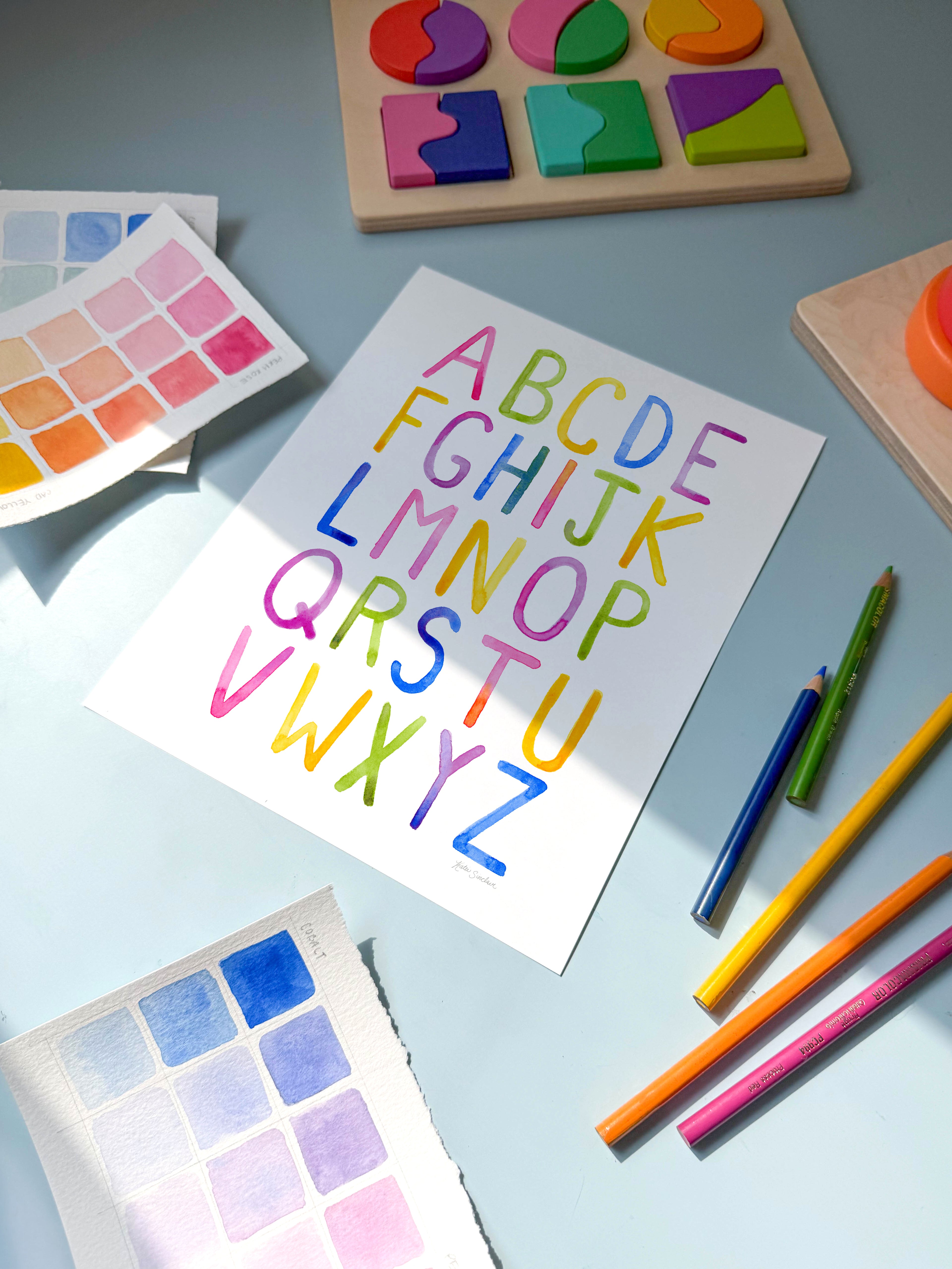 simple alphabet watercolor archival print by Katie Sinclair on a desk with colored pencils and a wooden puzzle.