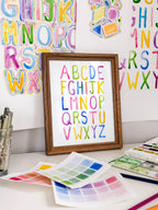 Framed watercolor simple alphabet archival print by Katie Sinclair on a desk with colorful letters on the wall