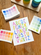 colorful watercolor simple alphabet archival print, surrounded by geometric toys on a wooden surface.  hand-painted by katie sinclair and perfect for a playroom or kid bedroom.