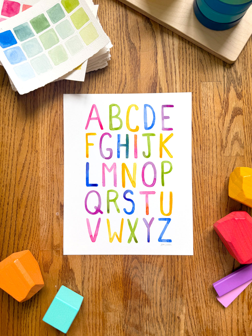 colorful watercolor simple alphabet archival print, surrounded by geometric toys on a wooden surface.  hand-painted by katie sinclair and perfect for a playroom or kid bedroom.