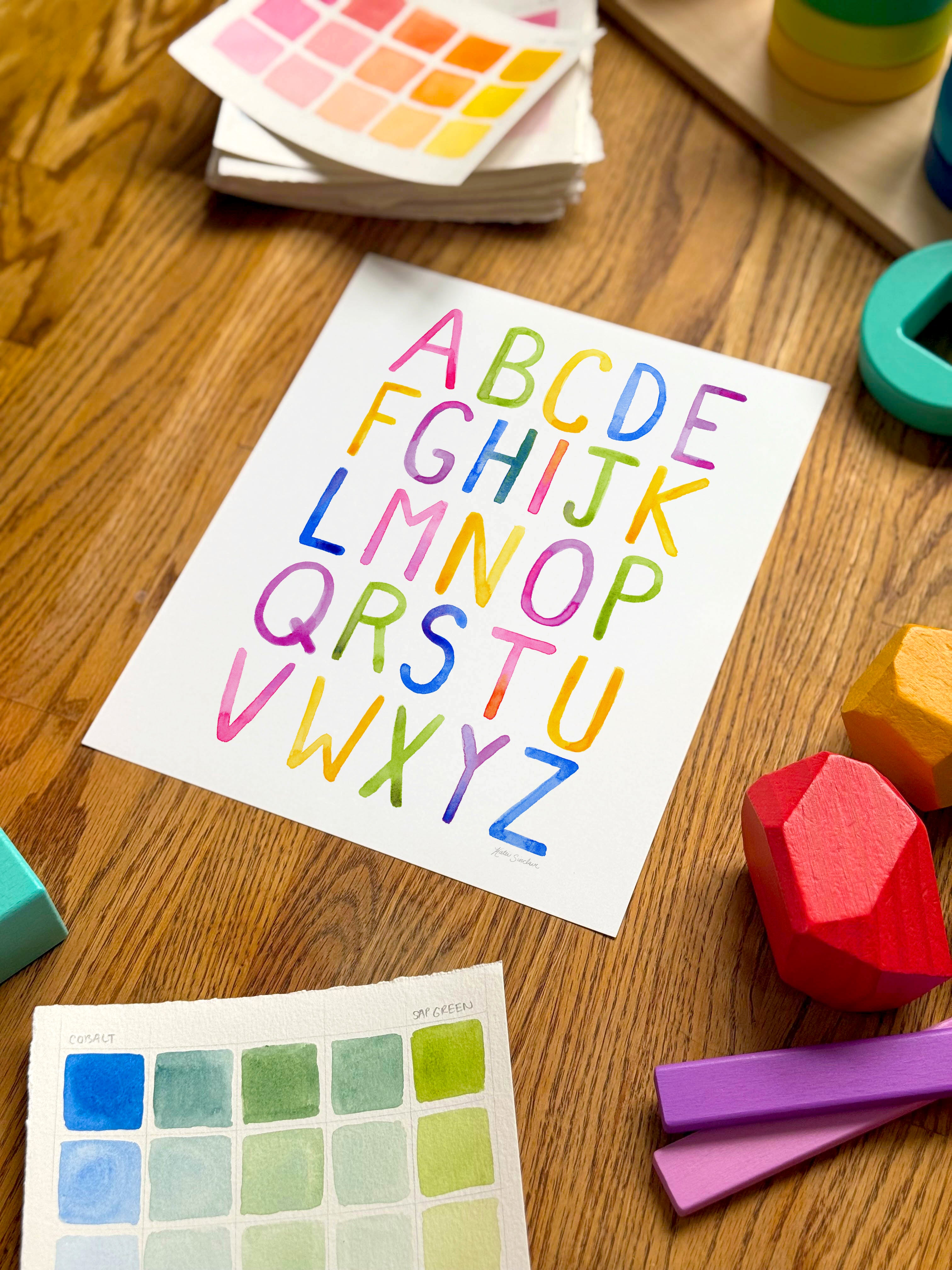colorful watercolor simple alphabet archival print, surrounded by geometric toys on a wooden surface.  hand-painted by katie sinclair and perfect for a playroom or kid bedroom.