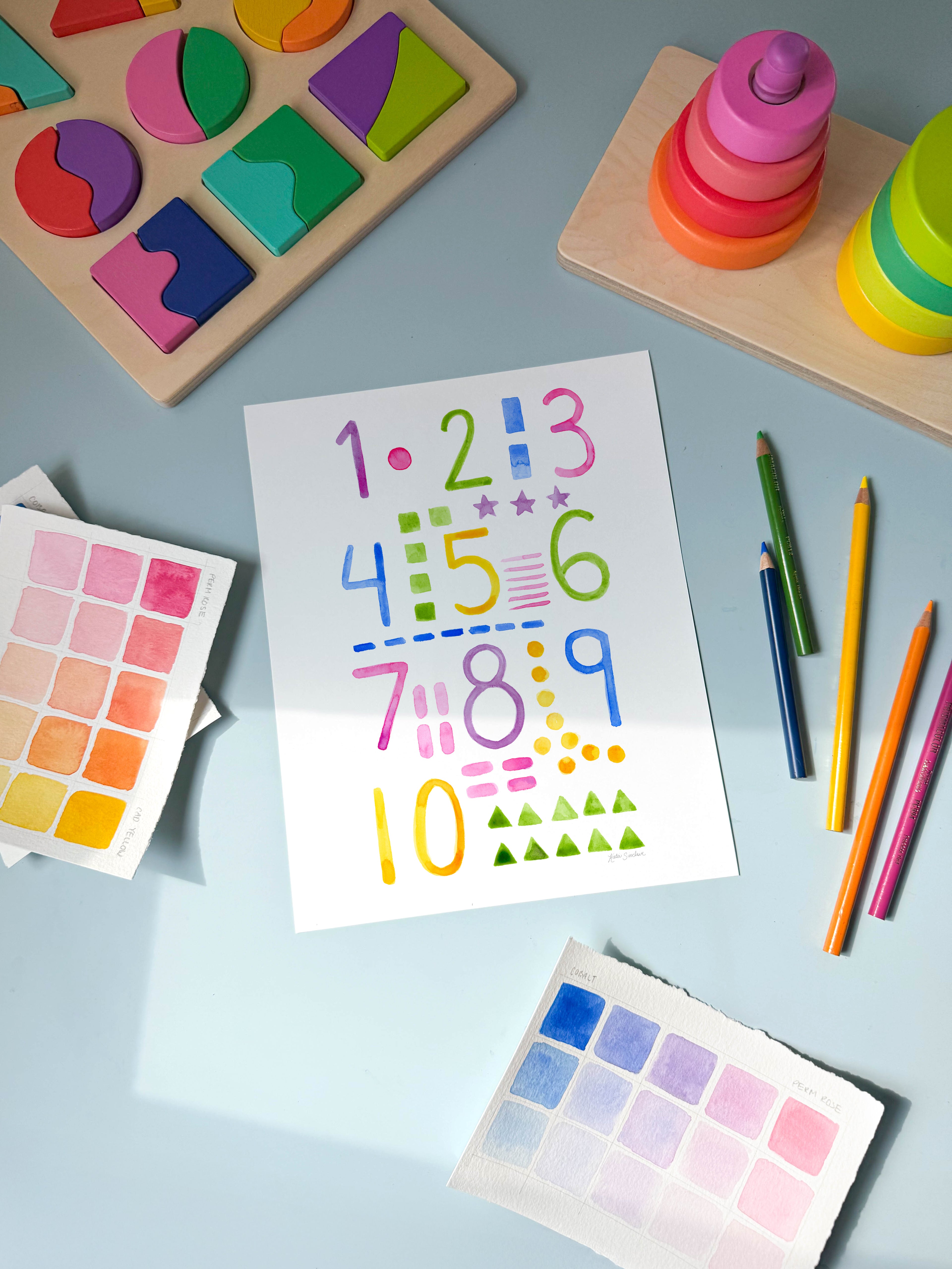 numbers and shapes watercolor archival print by Katie Sinclair on a desk with colored pencils and a wooden puzzle.