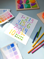 numbers and shapes watercolor archival print by Katie Sinclair on a desk with colored pencils and a wooden puzzle.
