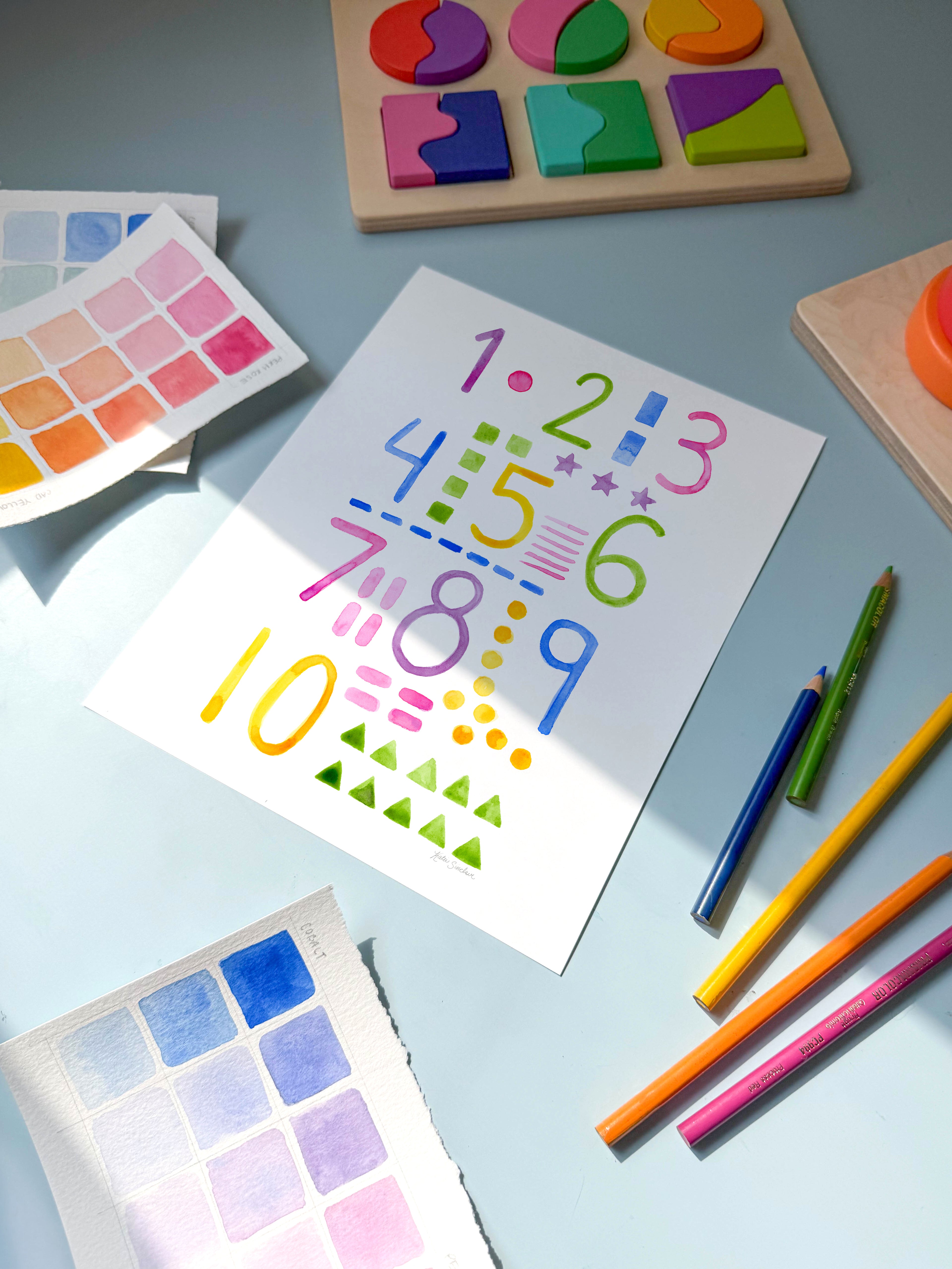 numbers and shapes watercolor archival print by Katie Sinclair on a desk with colored pencils and a wooden puzzle.