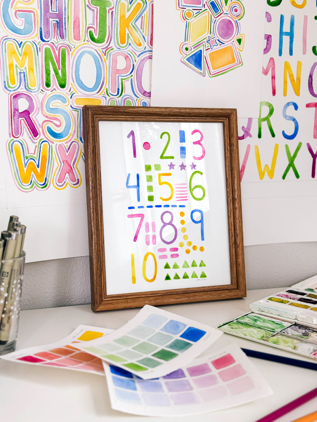 Framed watercolor numbers and shapes archival print by Katie Sinclair on a desk with colorful letters on the wall