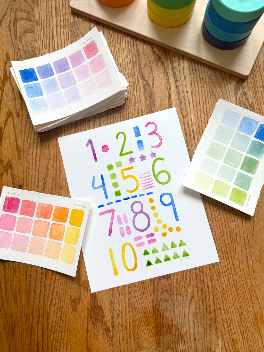 colorful watercolor numbers and shapes art print, surrounded by geometric toys on a wooden surface.  hand-painted by katie sinclair and perfect for a playroom or kid bedroom.