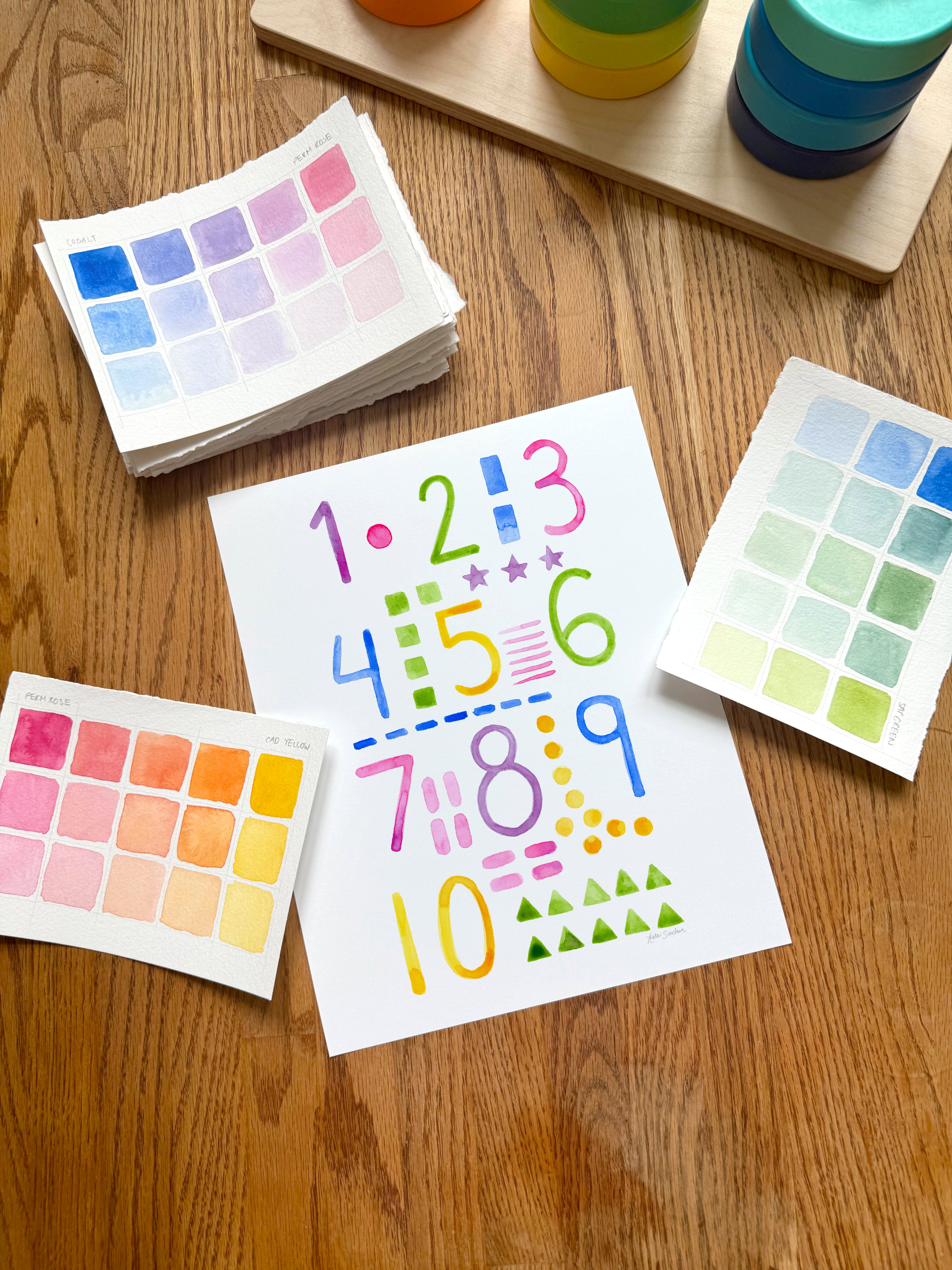 colorful watercolor numbers and shapes art print, surrounded by geometric toys on a wooden surface.  hand-painted by katie sinclair and perfect for a playroom or kid bedroom.
