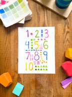 colorful watercolor numbers and shapes art print, surrounded by geometric toys on a wooden surface.  hand-painted by katie sinclair and perfect for a playroom or kid bedroom.