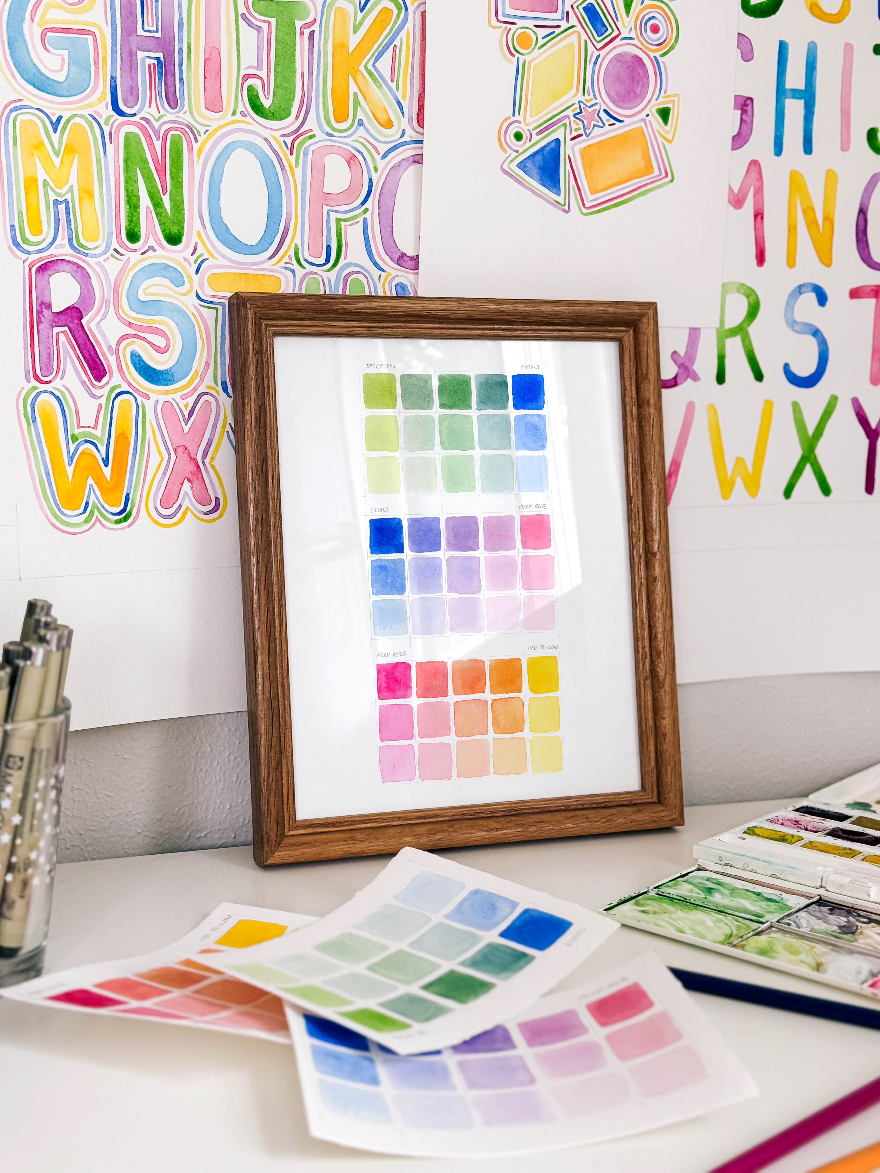 Framed watercolor color swatch chart archival print by Katie Sinclair on a desk with colorful letters on the wall