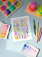 bubble letter alphabet watercolor archival print by Katie Sinclair on a desk with colored pencils and a wooden puzzle.