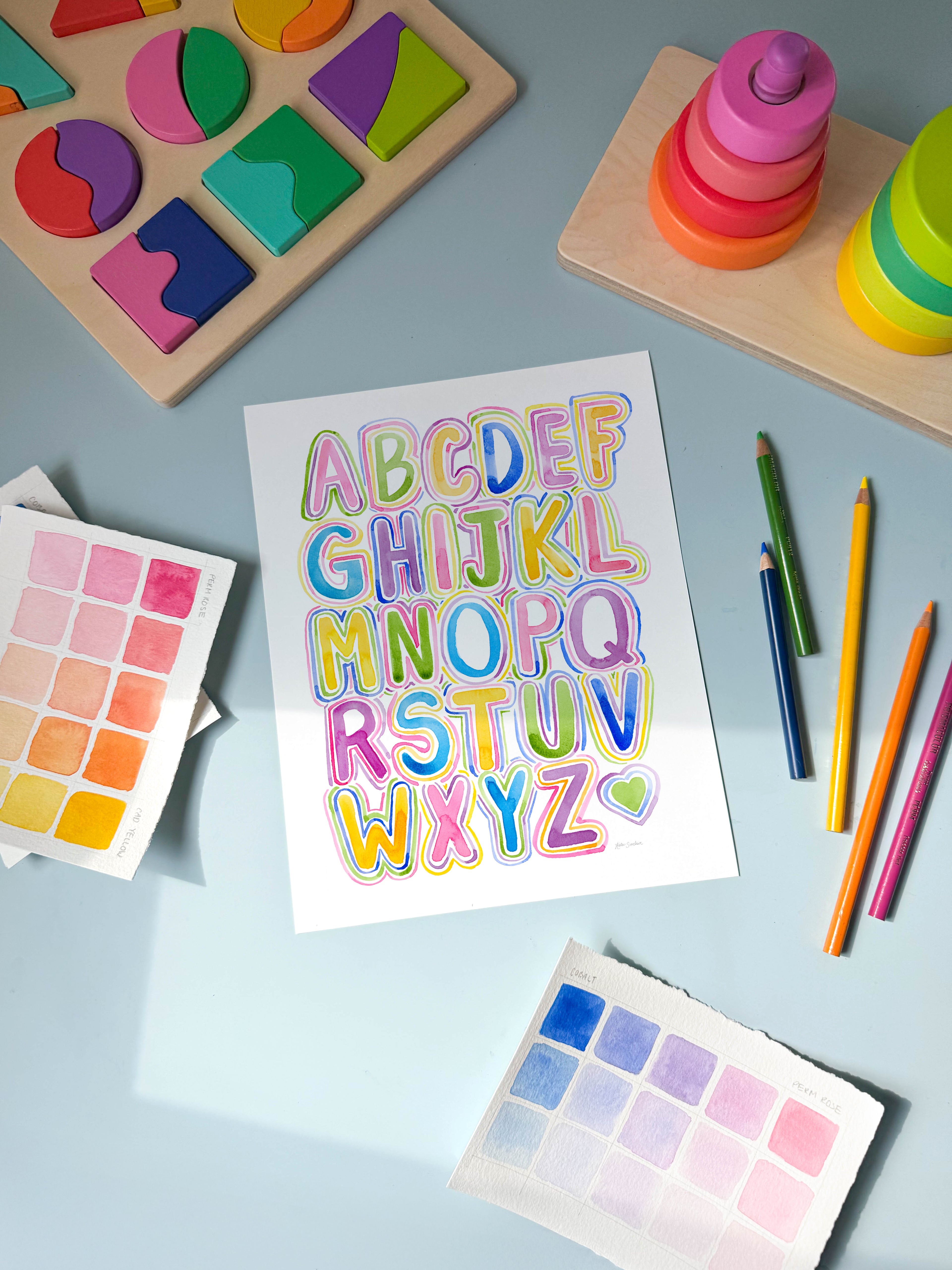bubble letter alphabet watercolor archival print by Katie Sinclair on a desk with colored pencils and a wooden puzzle.