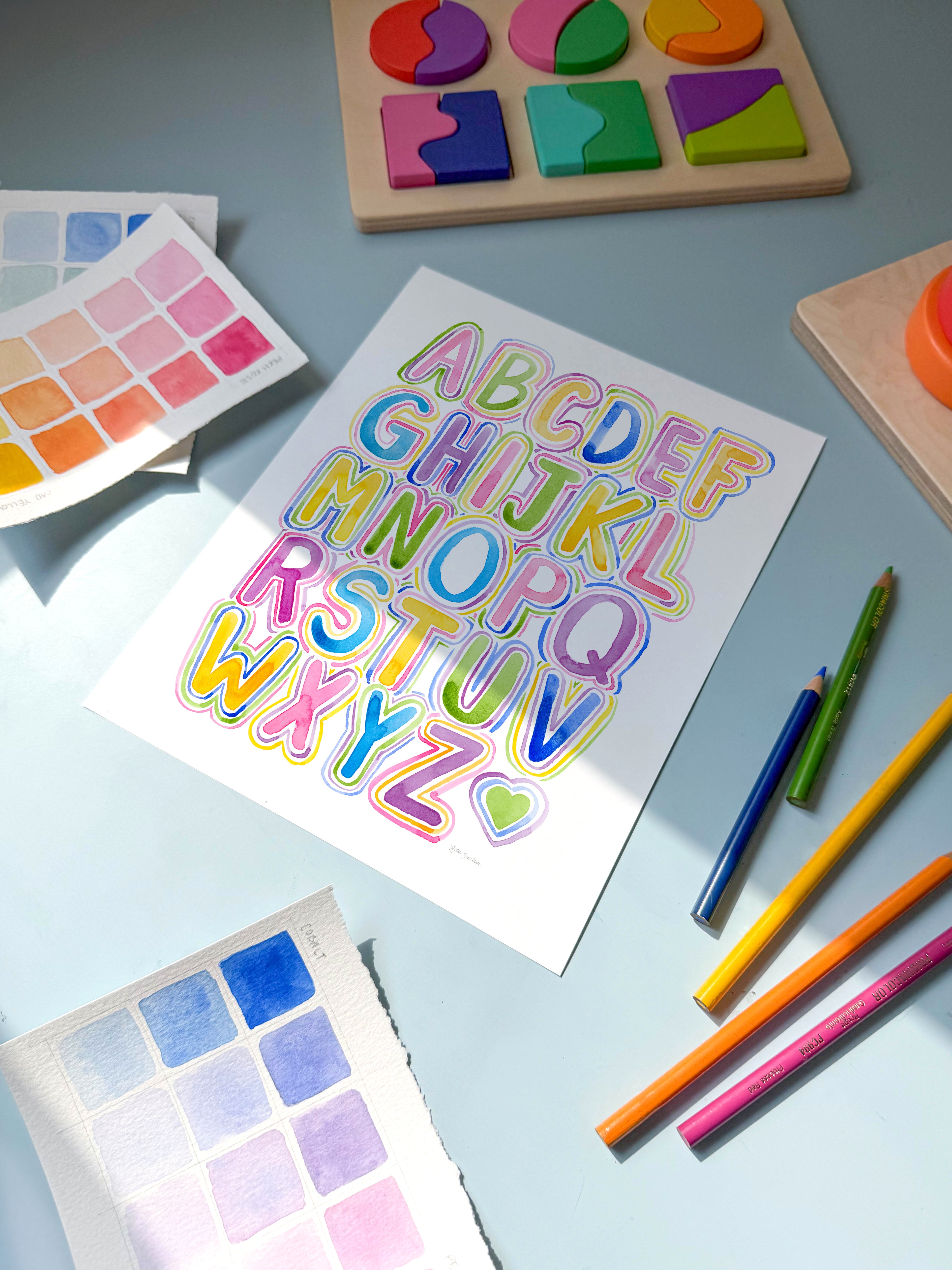 bubble letter alphabet watercolor archival print by Katie Sinclair on a desk with colored pencils and a wooden puzzle.