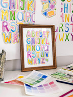 Framed watercolor bubble letter alphabet archival print by Katie Sinclair on a desk with colorful letters on the wall
