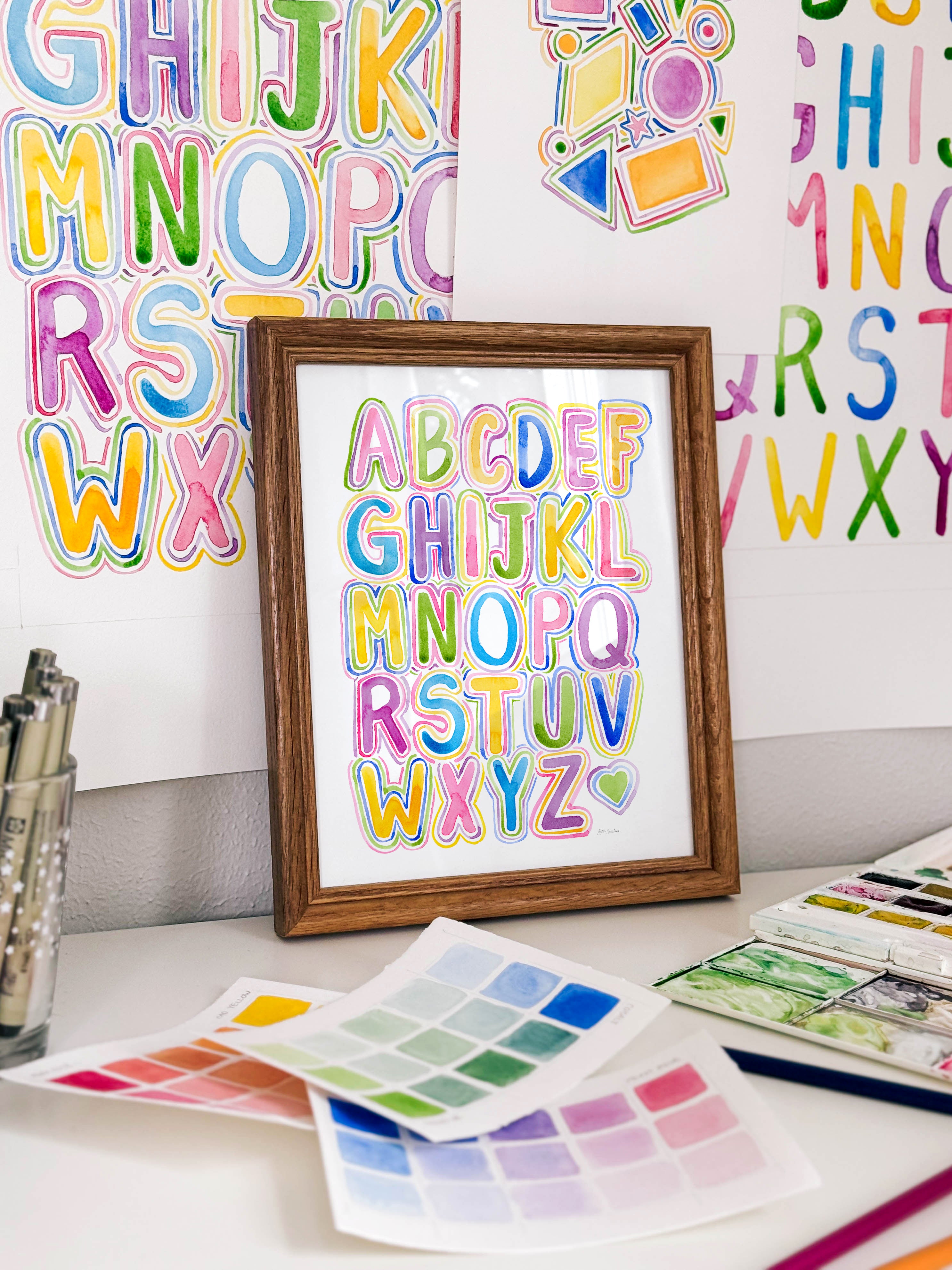 Framed watercolor bubble letter alphabet archival print by Katie Sinclair on a desk with colorful letters on the wall