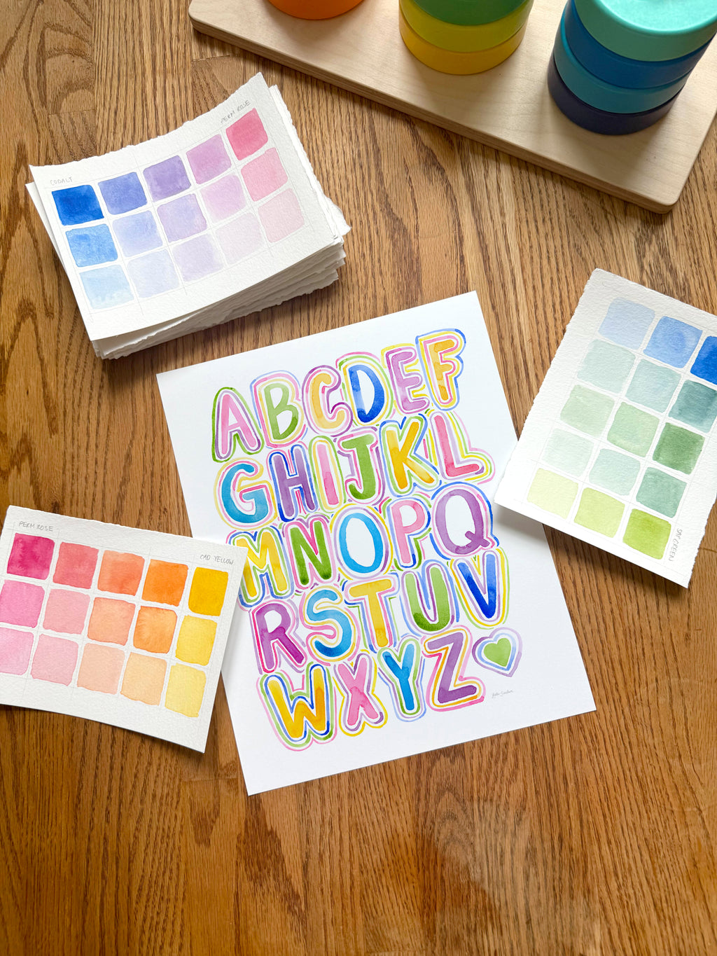 colorful alphabet watercolor art print, surrounded by geometric toys on a wooden surface.  hand-painted by katie sinclair and perfect for a playroom or kid bedroom.