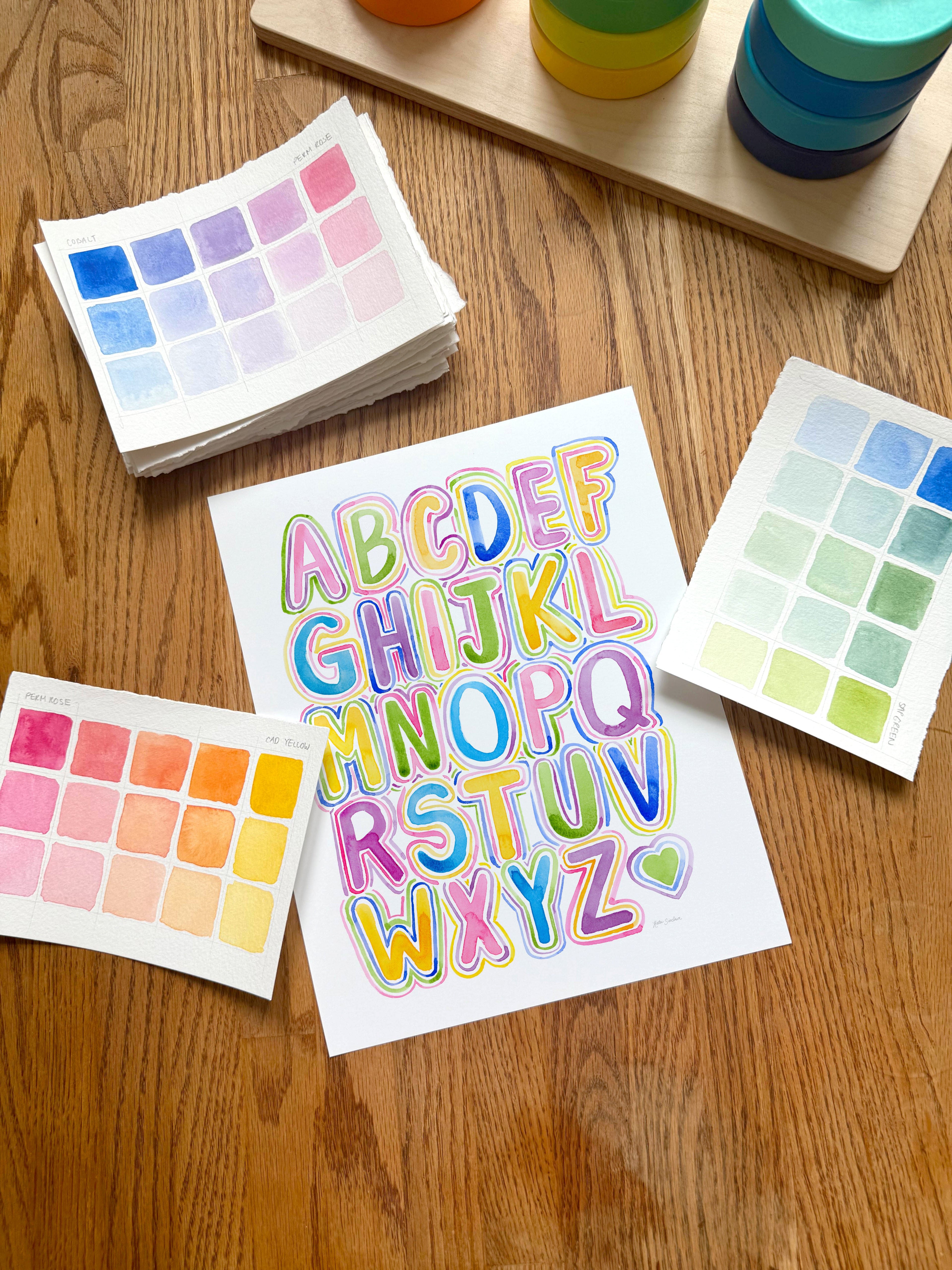 colorful alphabet watercolor art print, surrounded by geometric toys on a wooden surface.  hand-painted by katie sinclair and perfect for a playroom or kid bedroom.
