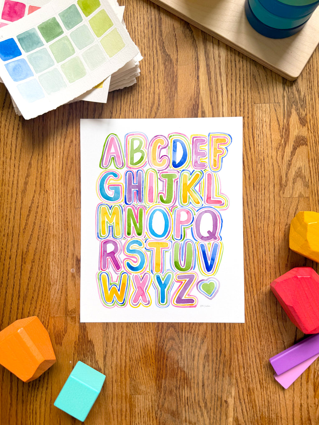 colorful alphabet watercolor art print, surrounded by geometric toys on a wooden surface.  hand-painted by katie sinclair and perfect for a playroom or kid bedroom.
