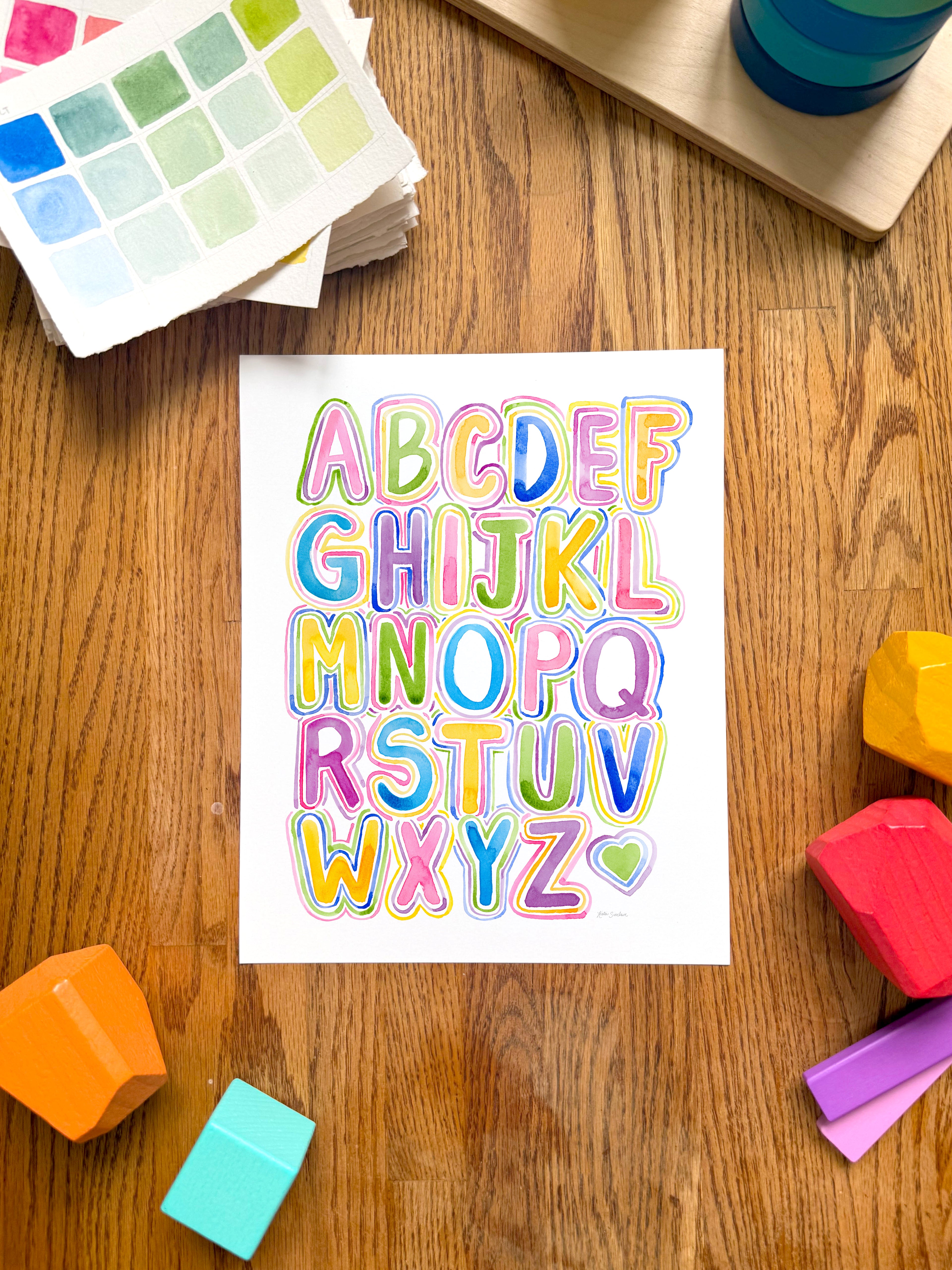 colorful alphabet watercolor art print, surrounded by geometric toys on a wooden surface.  hand-painted by katie sinclair and perfect for a playroom or kid bedroom.