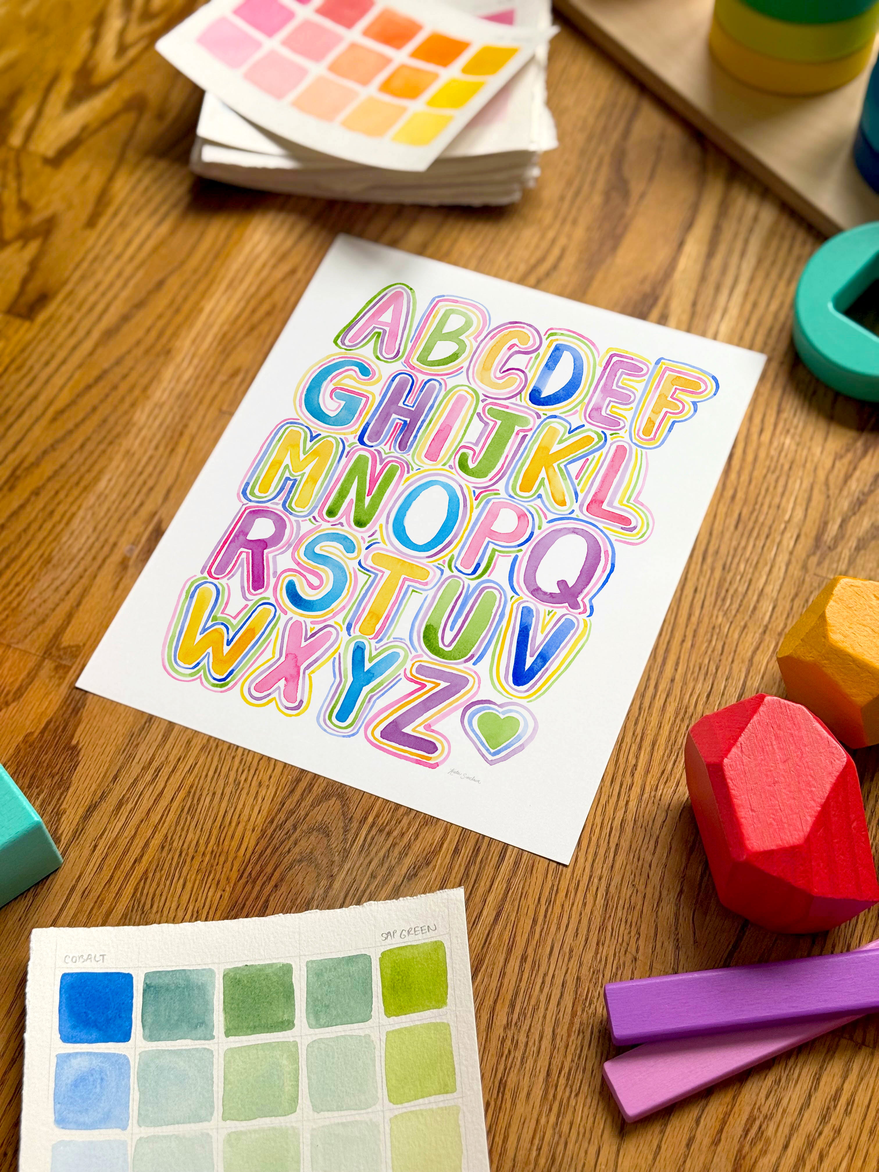 colorful alphabet watercolor art print, surrounded by geometric toys on a wooden surface.  hand-painted by katie sinclair and perfect for a playroom or kid bedroom.