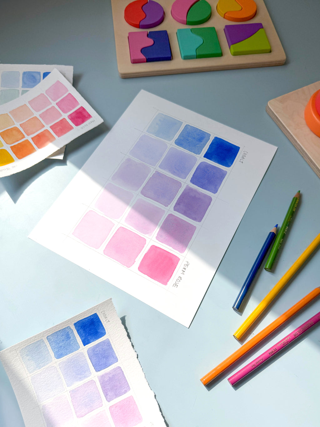 pink and blue color swatch watercolor archival print by Katie Sinclair on a desk with colored pencils and a wooden puzzle.