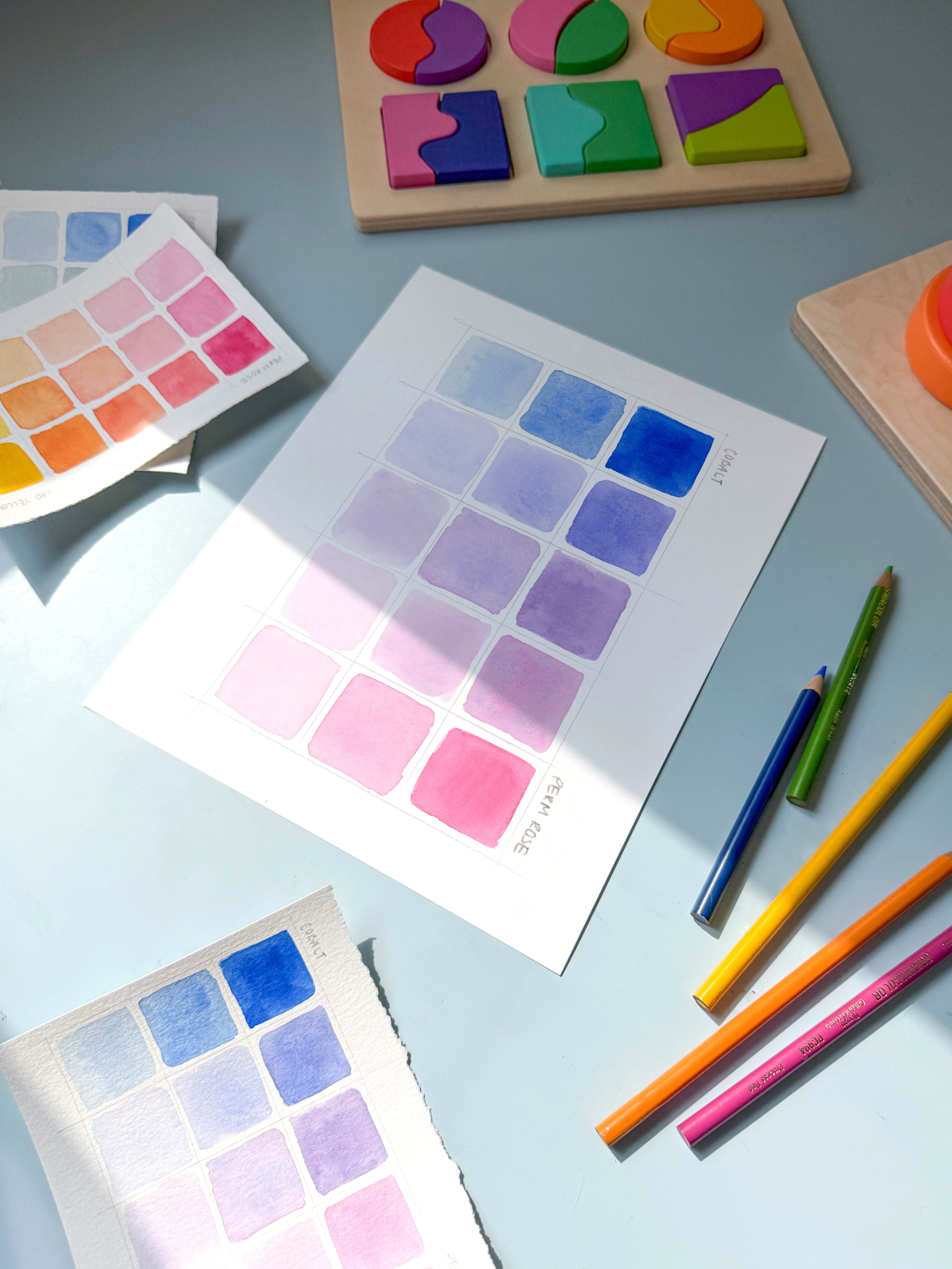 pink and blue color swatch watercolor archival print by Katie Sinclair on a desk with colored pencils and a wooden puzzle.