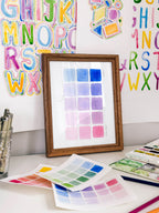 Framed watercolor color swatch chart archival print by Katie Sinclair on a desk with colorful letters on the wall