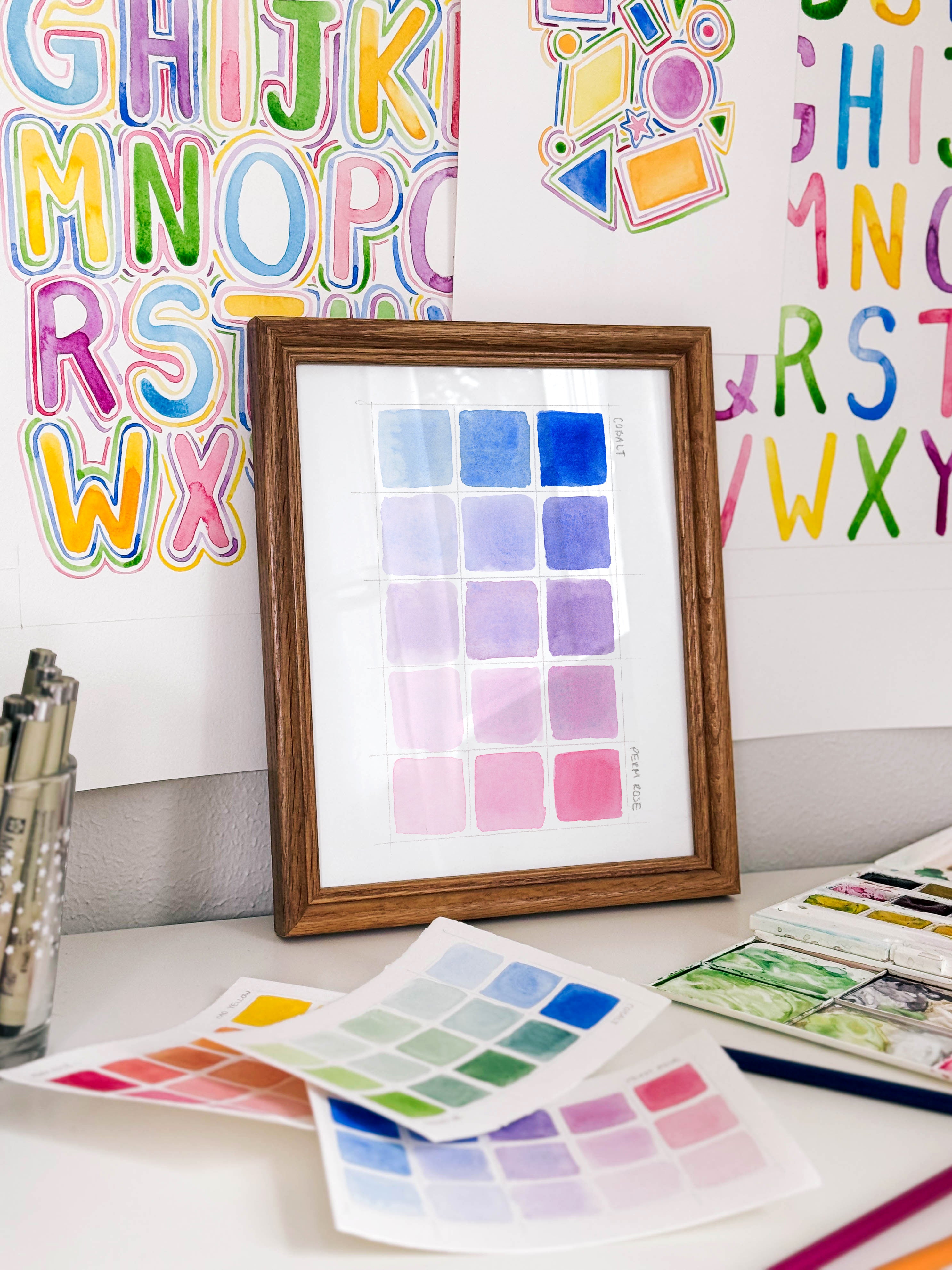 Framed watercolor color swatch chart archival print by Katie Sinclair on a desk with colorful letters on the wall