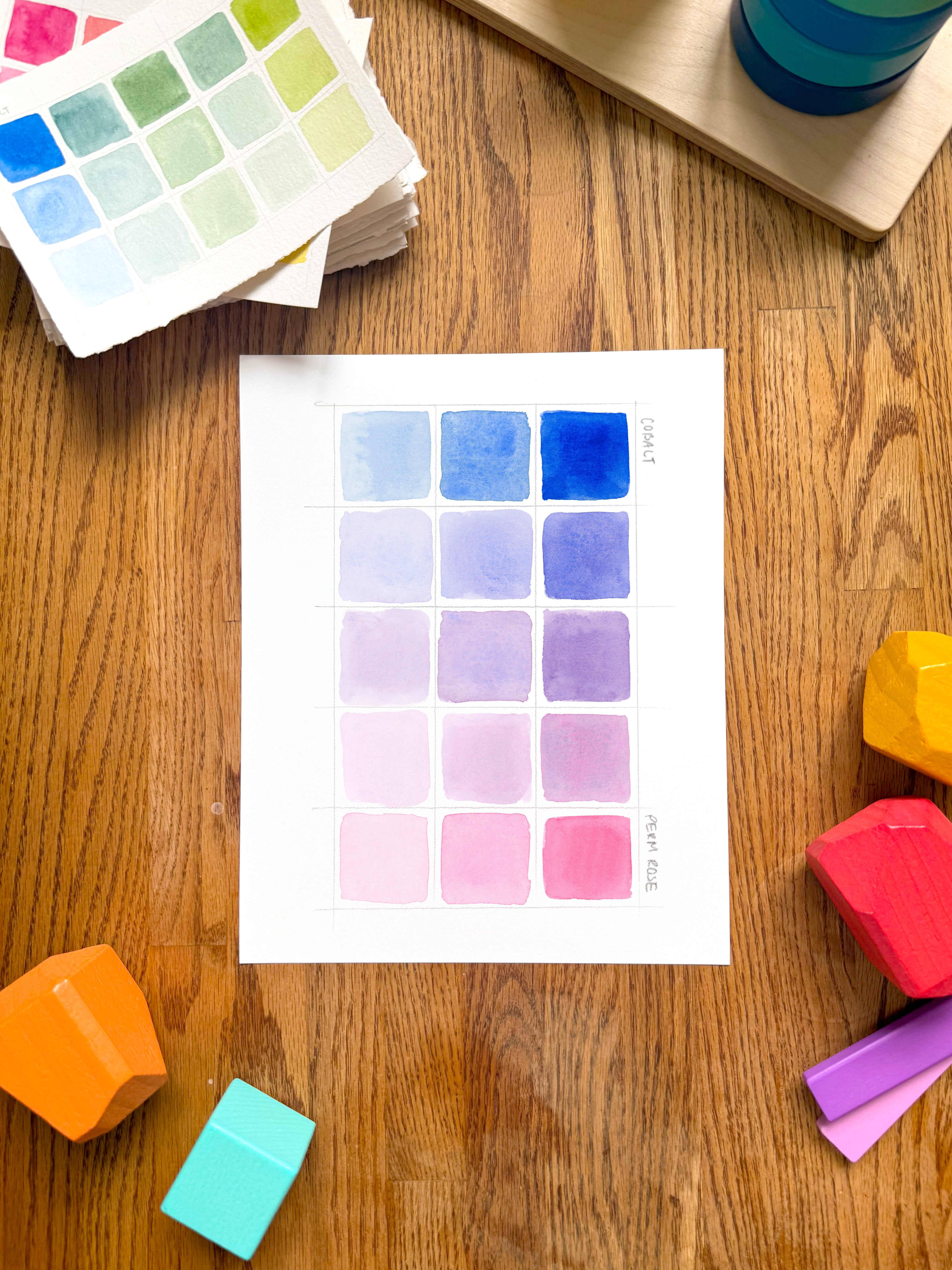 pink and blue color swatch watercolor art print, surrounded by geometric toys on a wooden surface