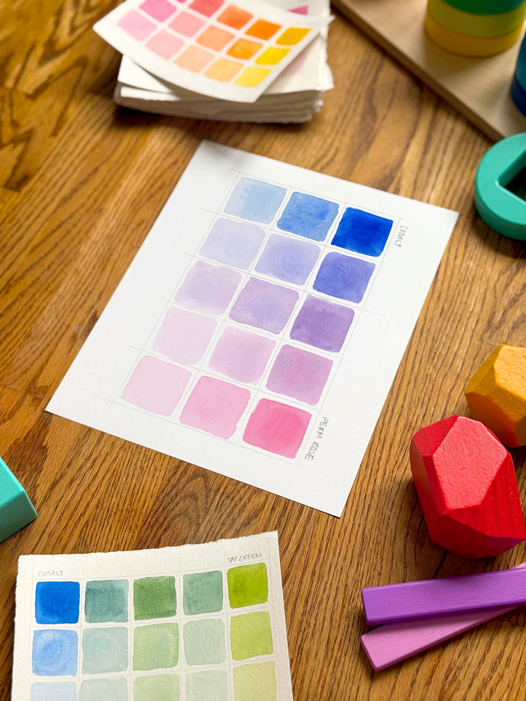 pink and blue color swatch watercolor art print, surrounded by geometric toys on a wooden surface