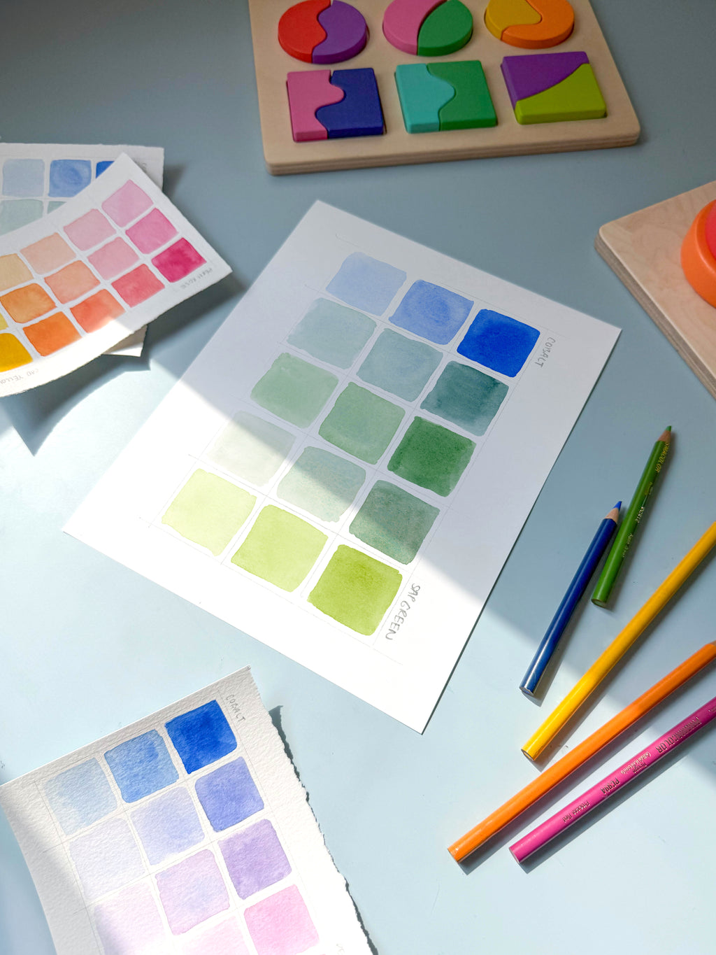 blue and green color swatch watercolor archival print by Katie Sinclair on a desk with colored pencils and a wooden puzzle.