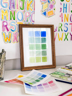 Framed watercolor color swatch chart archival print by Katie Sinclair with blue and green on a desk with colorful letters on the wall