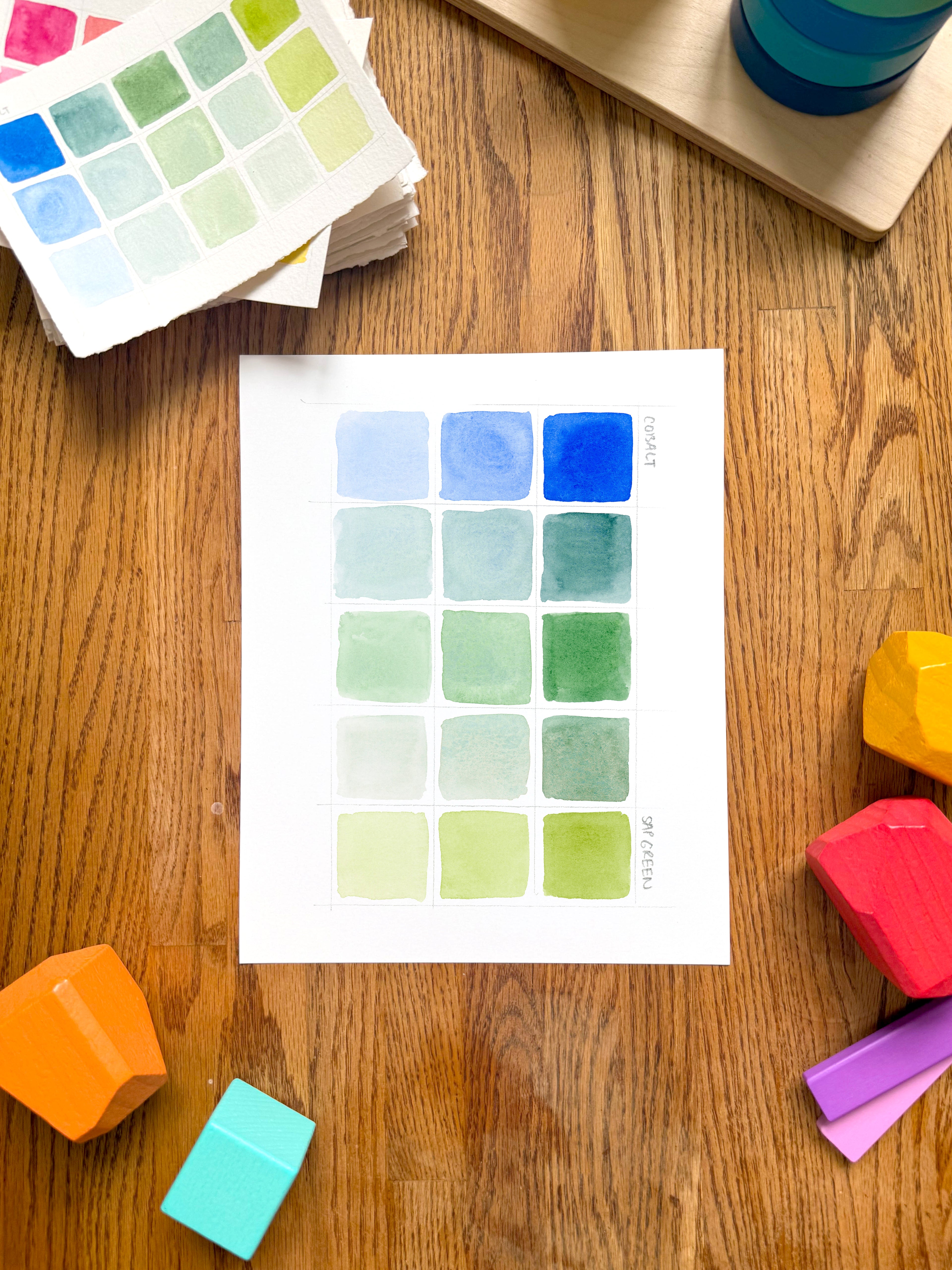 blue and green color swatch watercolor art print, surrounded by geometric toys on a wooden surface