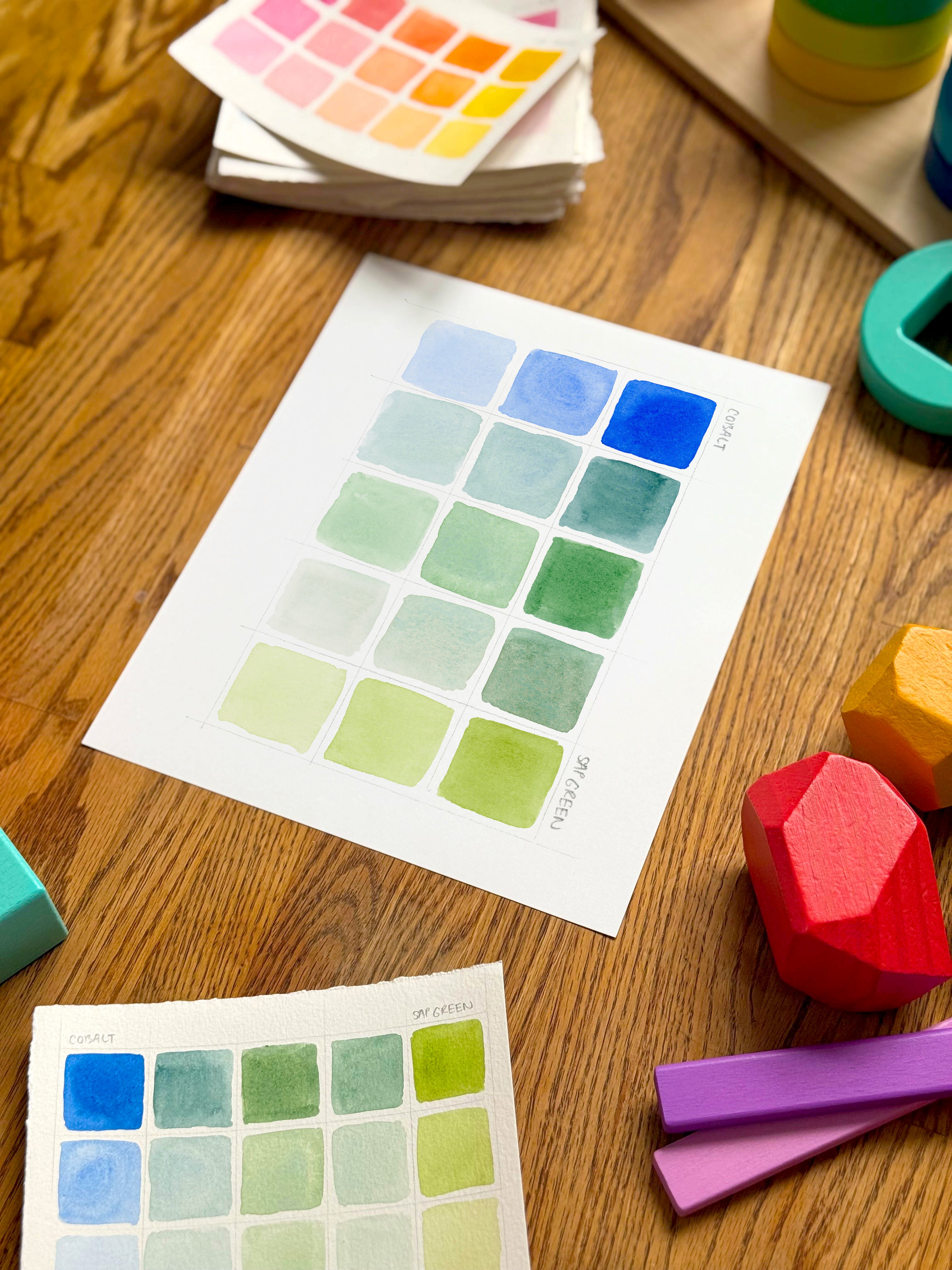 blue and green color swatch watercolor art print, surrounded by geometric toys on a wooden surface