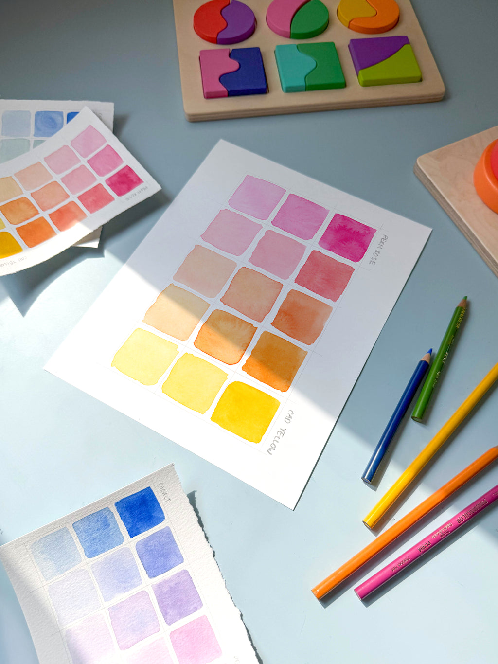 pink and yellow color swatch watercolor archival print by Katie Sinclair on a desk with colored pencils and a wooden puzzle.