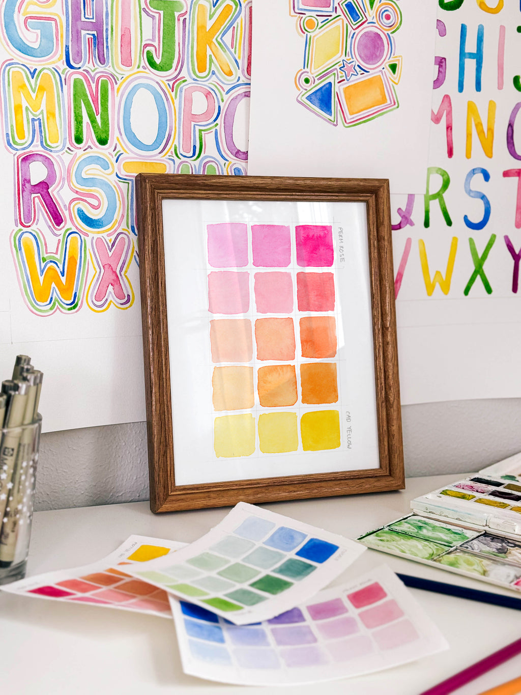 Framed watercolor color swatch chart archival print by Katie Sinclair on a desk with colorful letters on the wall