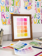 Framed watercolor color swatch chart archival print by Katie Sinclair on a desk with colorful letters on the wall