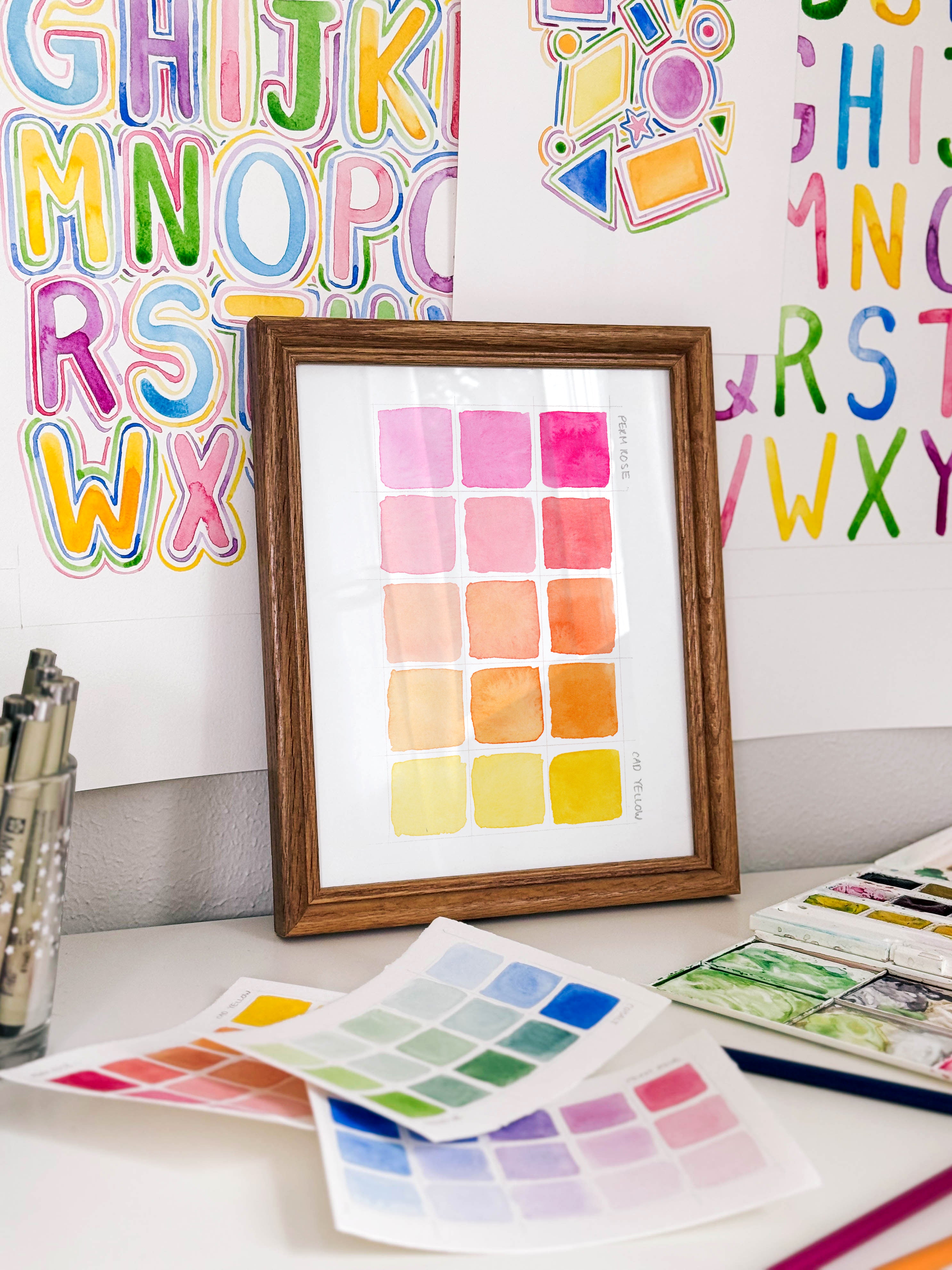 Framed watercolor color swatch chart archival print by Katie Sinclair on a desk with colorful letters on the wall