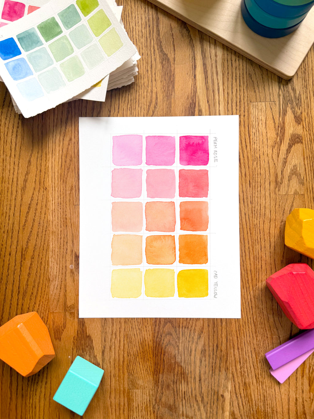 pink and yellow color swatch watercolor art print, surrounded by geometric toys on a wooden surface