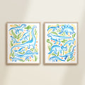 Two framed colorful blue, green, and yellow watercolor dinosaur-themed artworks on a beige wall, featuring a brontosaurus, t-rex, triceratops, raptor, spinosaurus, and plants.