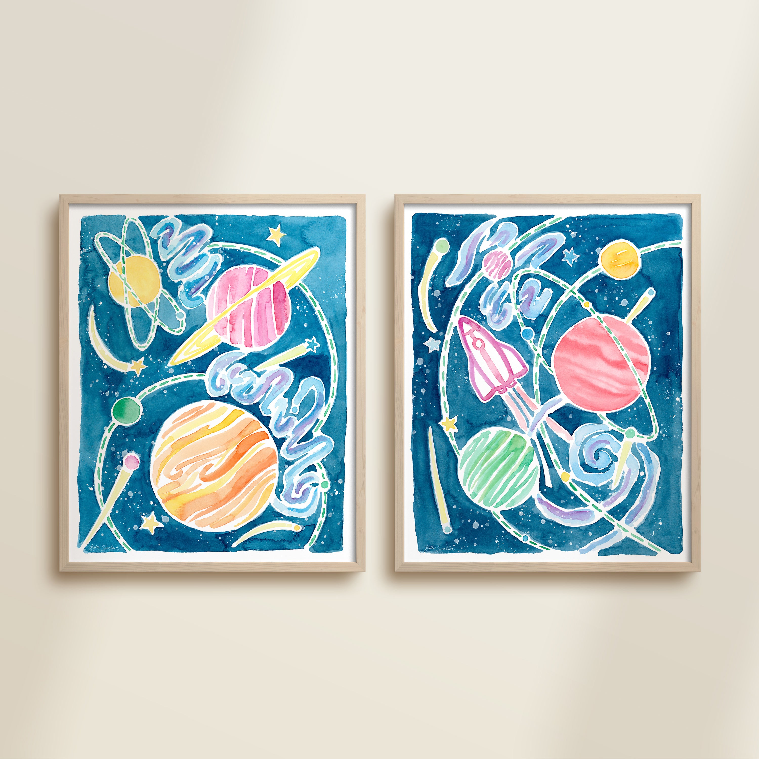 Two framed clolorful watercolor space-themed artworks on a beige wall, featuring planets, spaceships, stars, and a galaxy.