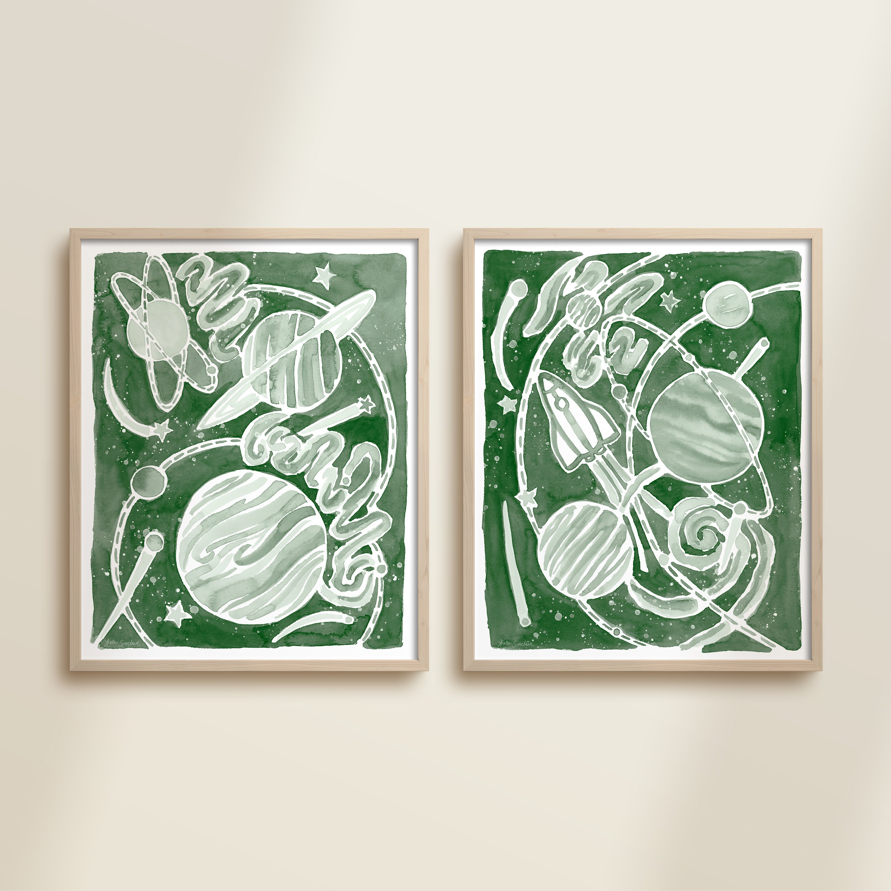 Two framed green watercolor space-themed artworks on a beige wall, featuring planets, spaceships, stars, and a galaxy.